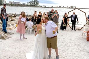 Beach wedding ceremony with guests gathered around couple under wooden arch.