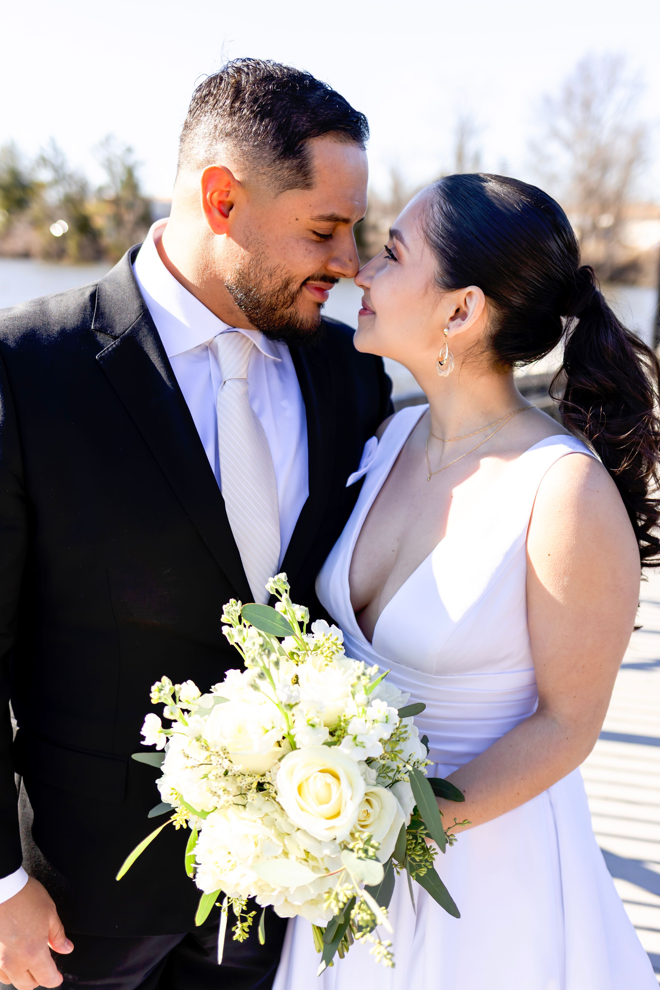 Newlywed couple shares intimate moment outdoors with elegant white rose bouquet on sunny winter day.