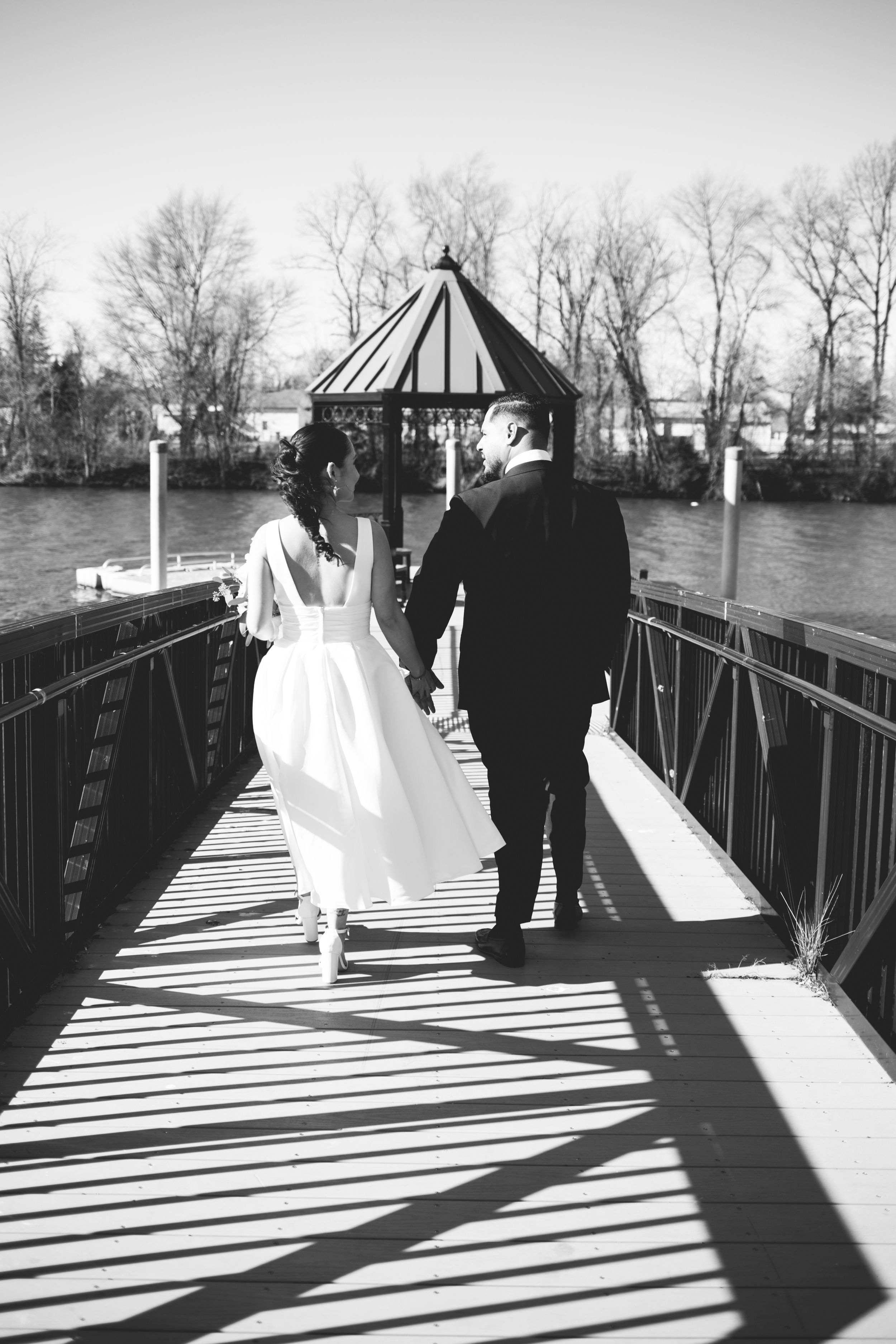 A couple in wedding attire walking across a dock with a gazebo, creating dramatic shadows in black and white photography.