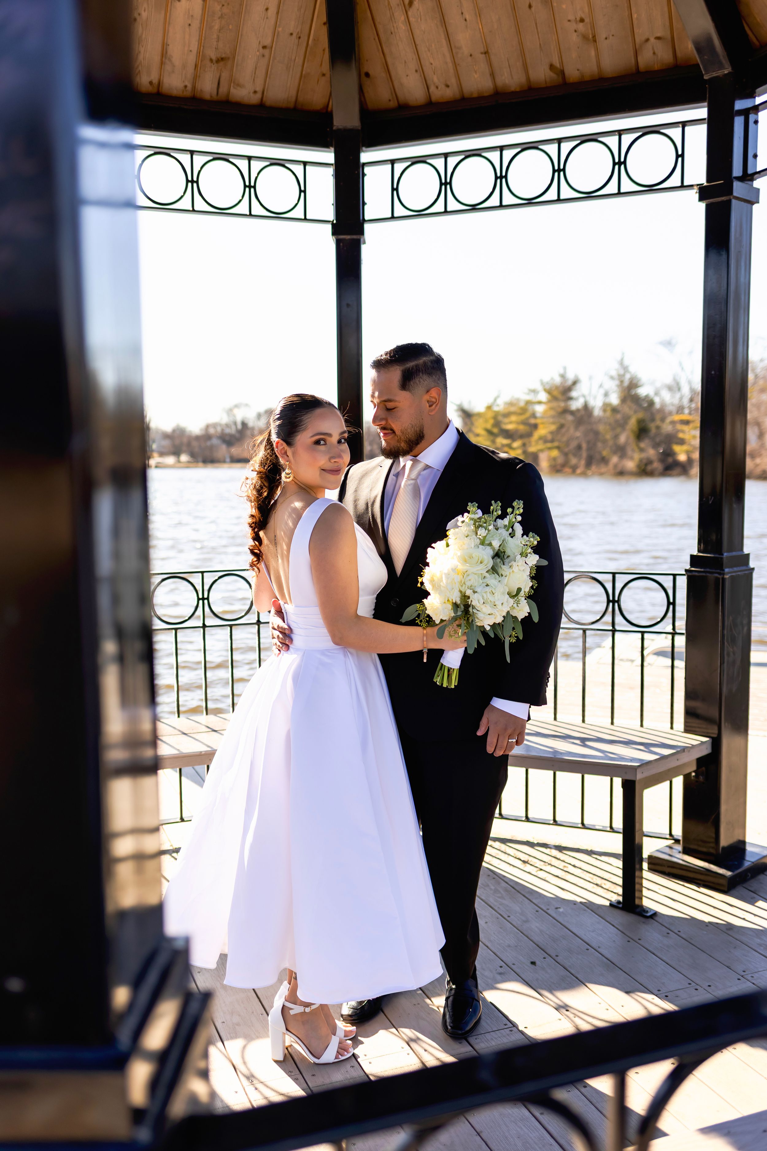 A wedding couple shares an intimate moment in a gazebo overlooking a lake while holding a white floral bouquet.