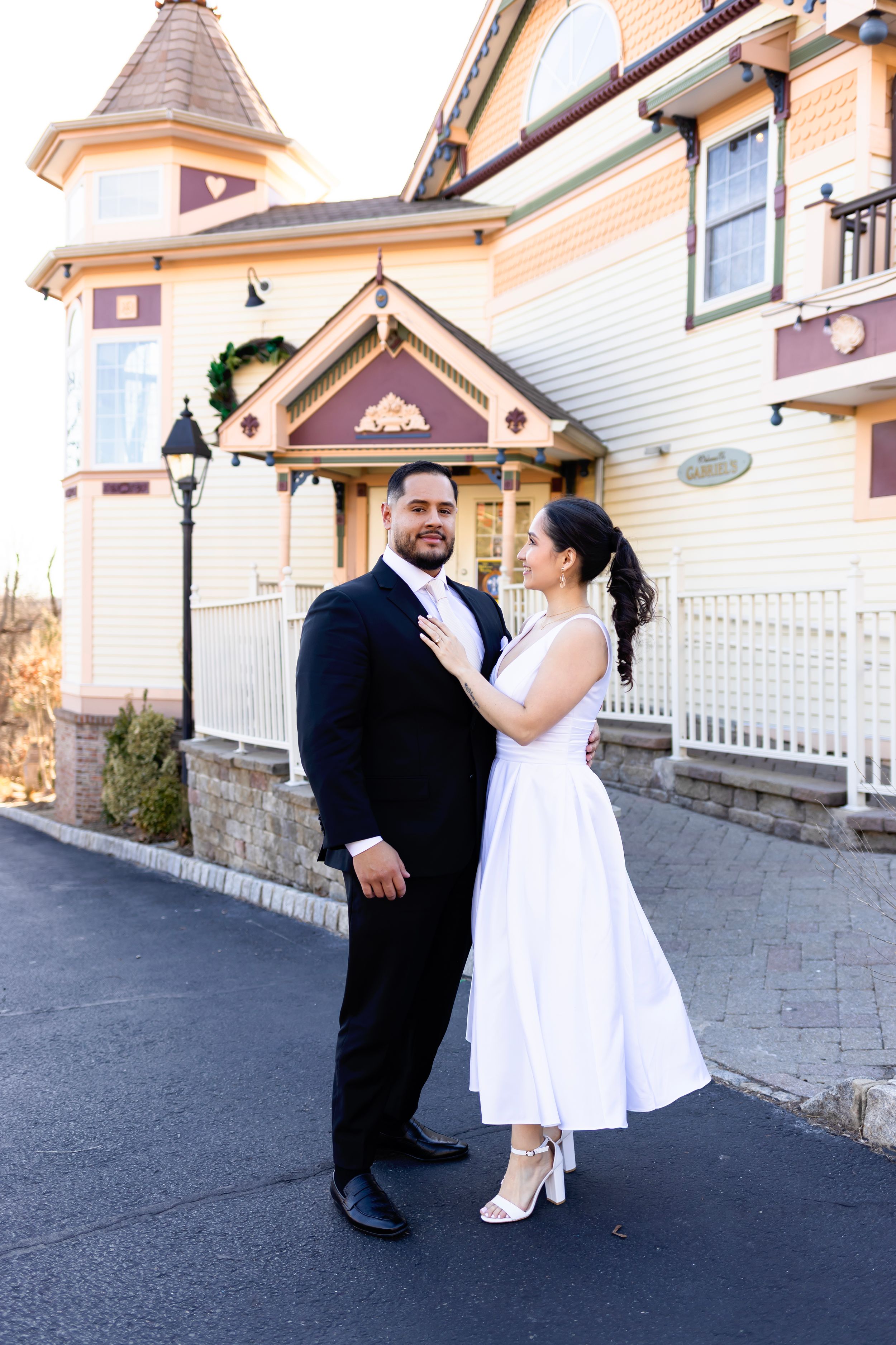A couple stands outside a charming Victorian-style house with cream colored walls and a turret on their wedding day.