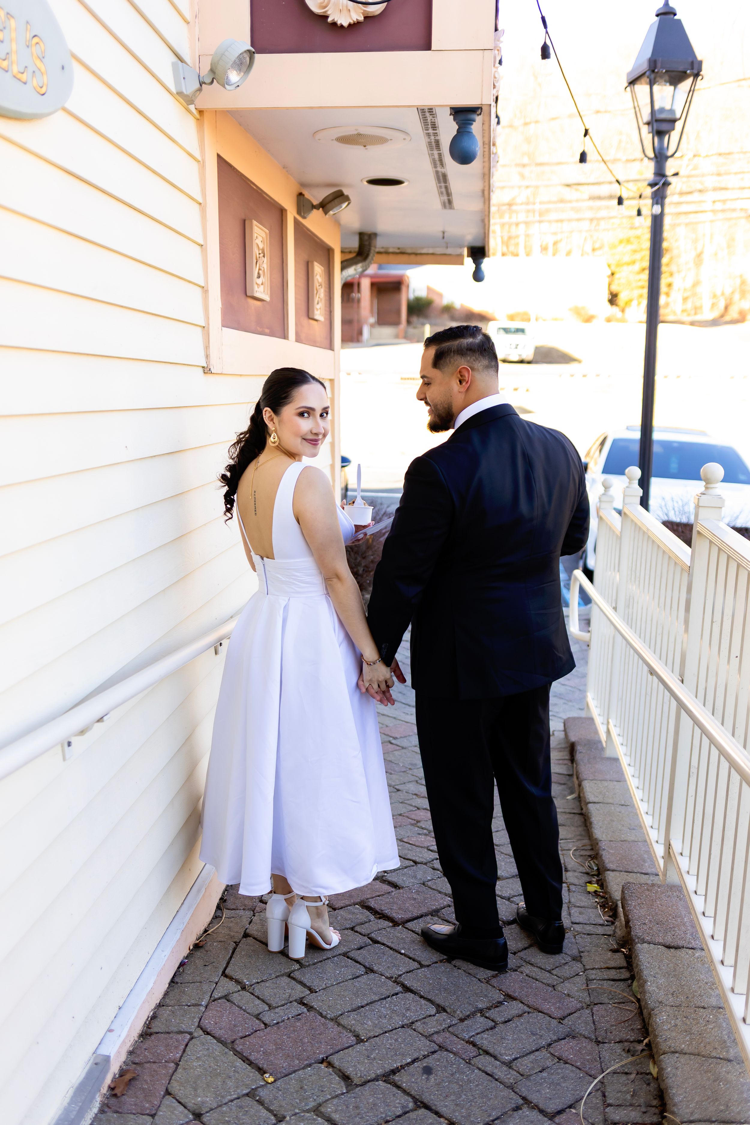 A couple in formal wedding attire walks along a brick paved covered walkway next to a white building with string lights above.