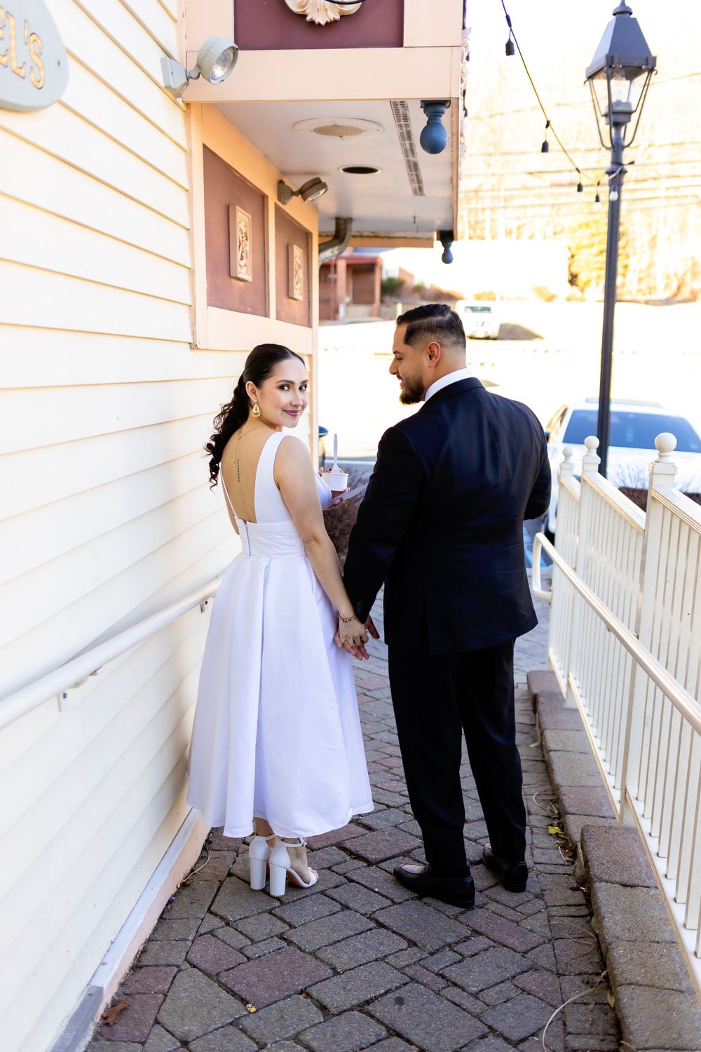 A couple in formal wedding attire walks along a brick paved covered walkway next to a white building with string lights above.