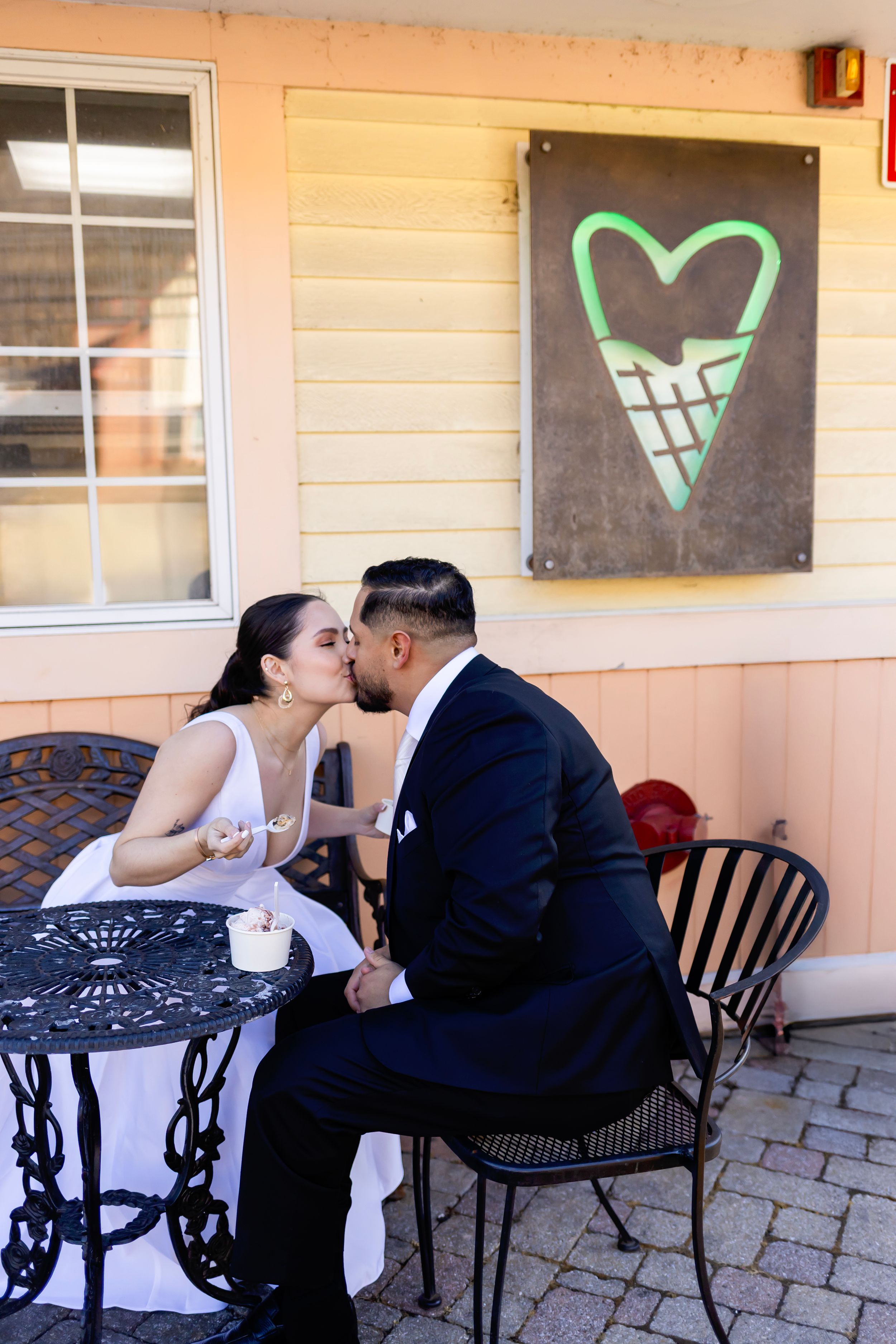 A couple shares a romantic moment at an outdoor cafe table beneath a heart-shaped wall art in green.