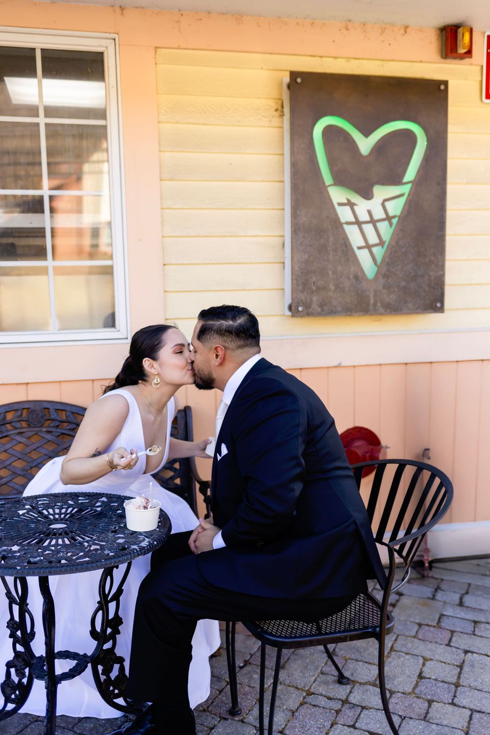 A couple shares a romantic moment at an outdoor cafe table beneath a heart-shaped wall art in green.