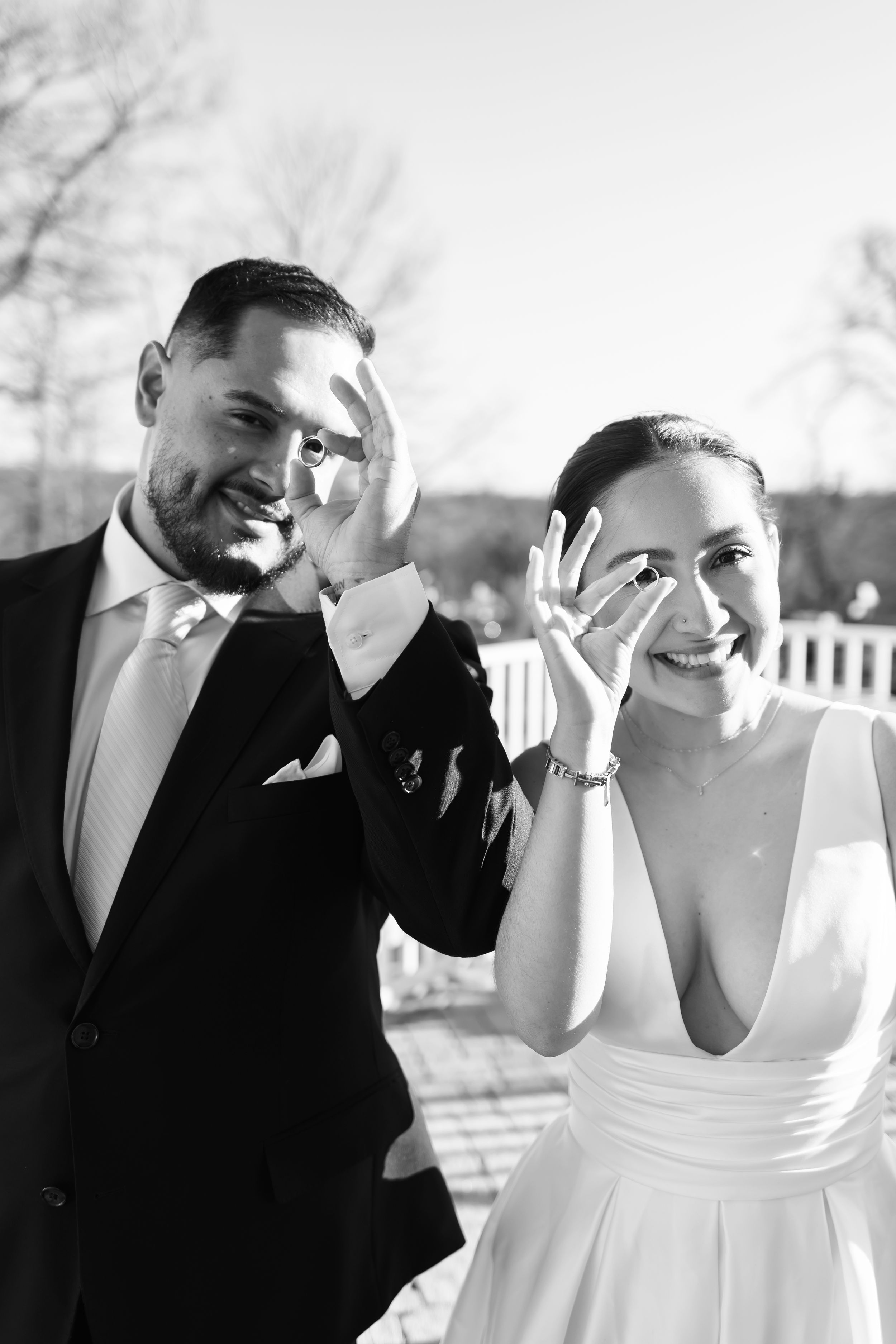 A joyful black and white photo of newlyweds showing off their rings in an outdoor setting.