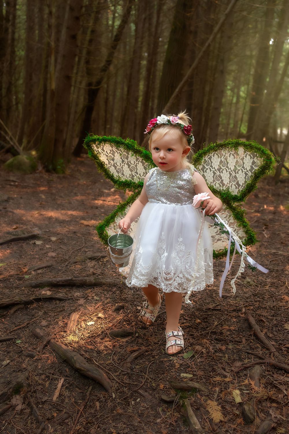 A child in a white lace dress and sparkly green fairy wings explores a magical forest setting.