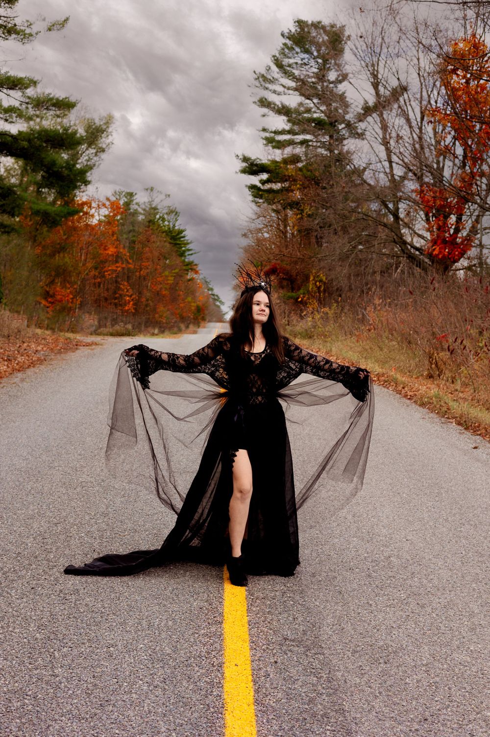 A figure in a flowing black dress poses dramatically on an autumn road with yellow lines and fall foliage.
