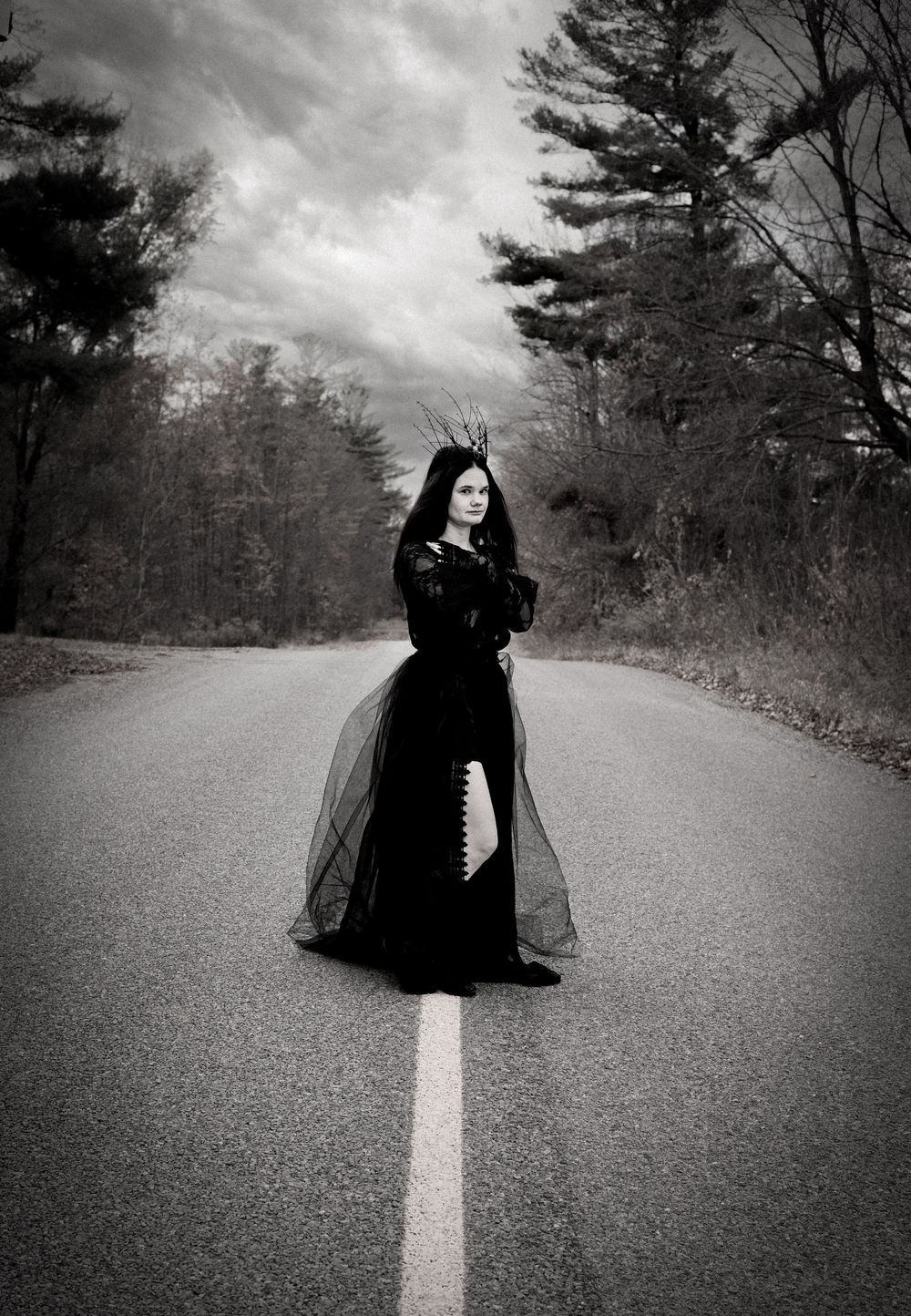 A gothic figure in a flowing black dress stands on an empty road in the woods under dramatic cloudy skies.
