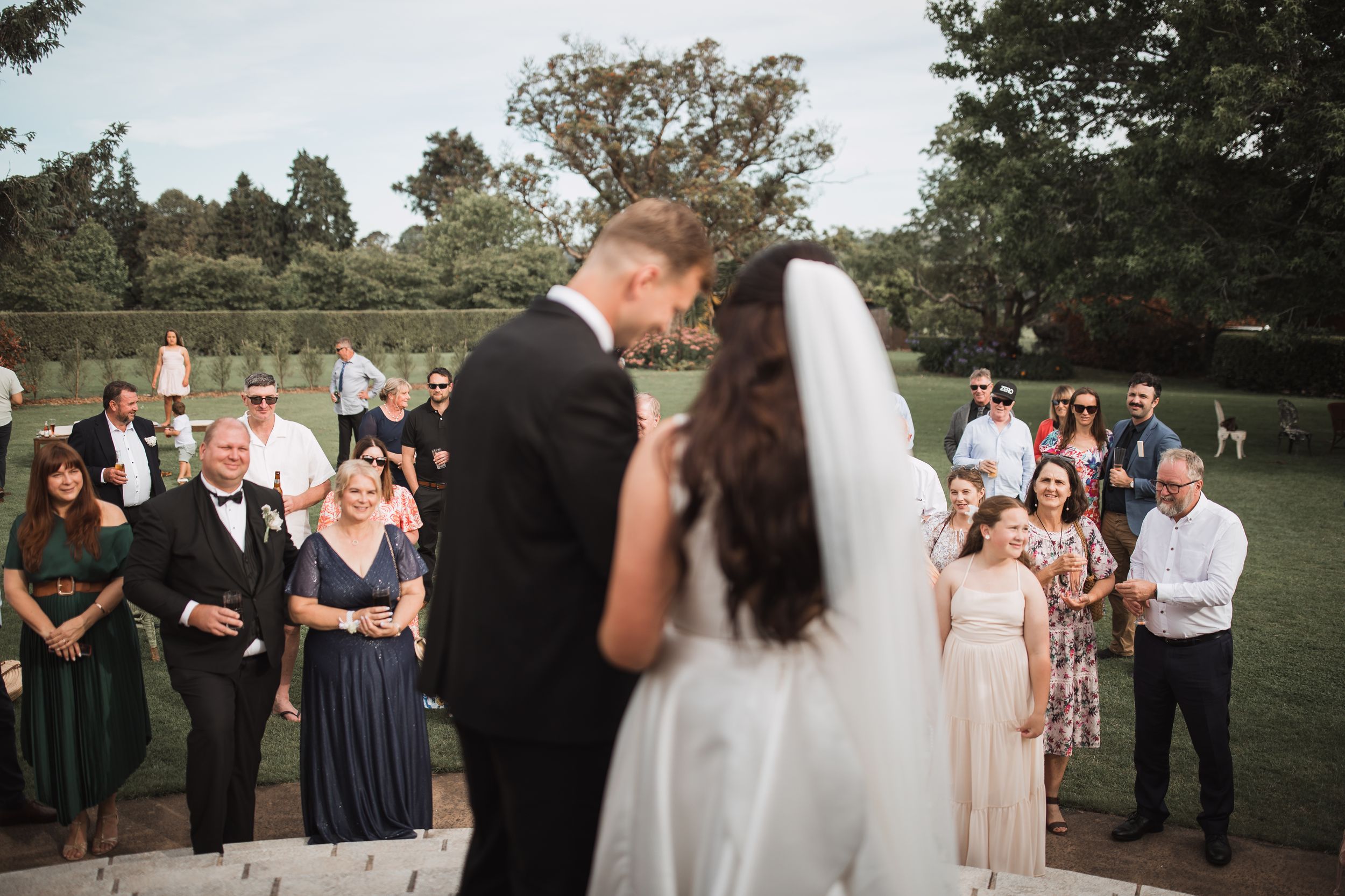 A wedding ceremony takes place outdoors with guests gathered in formal attire and floral decorations.