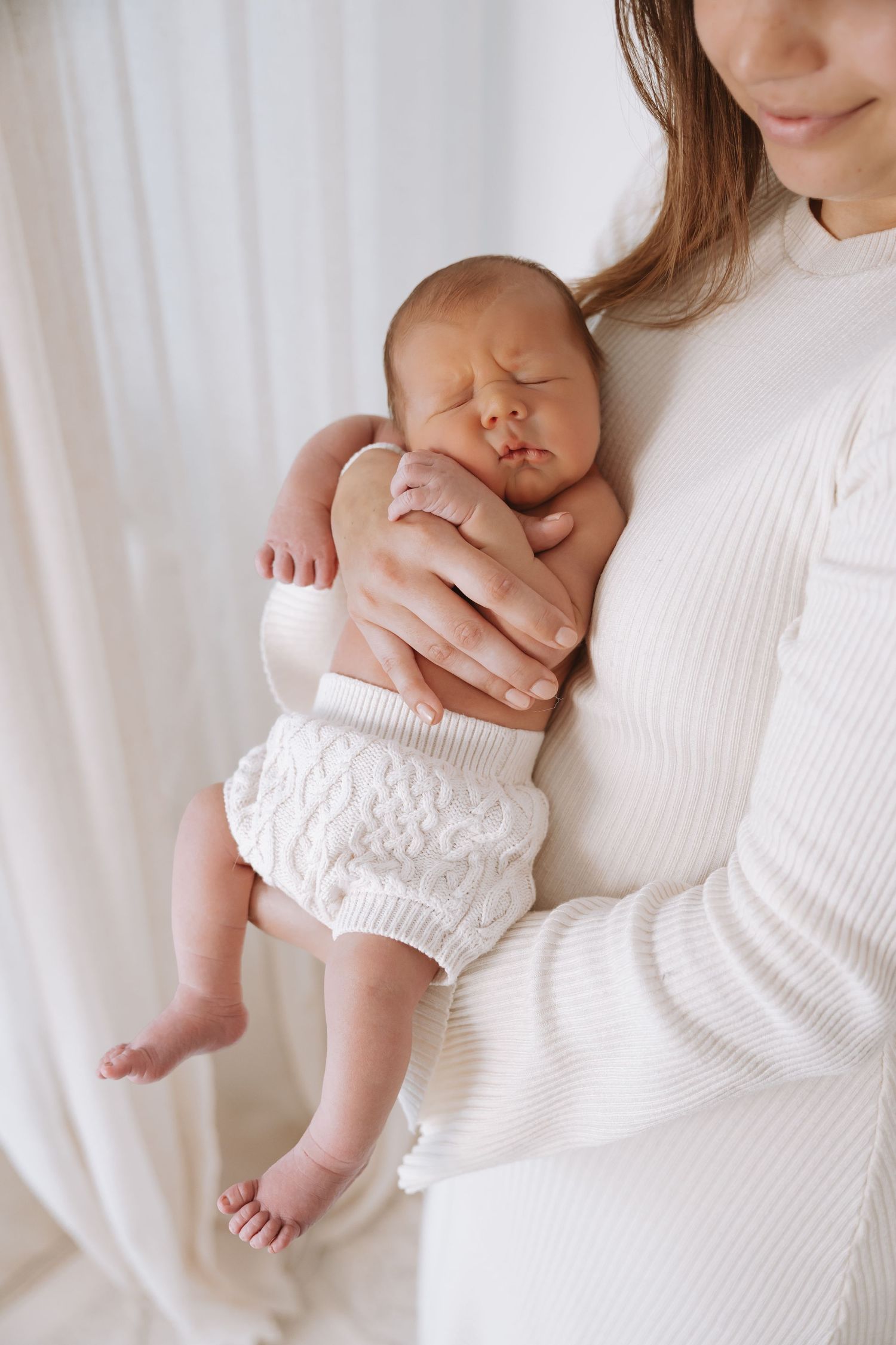 A newborn baby in white knit bloomers sleeps peacefully while being cradled against someone wearing a white sweater.