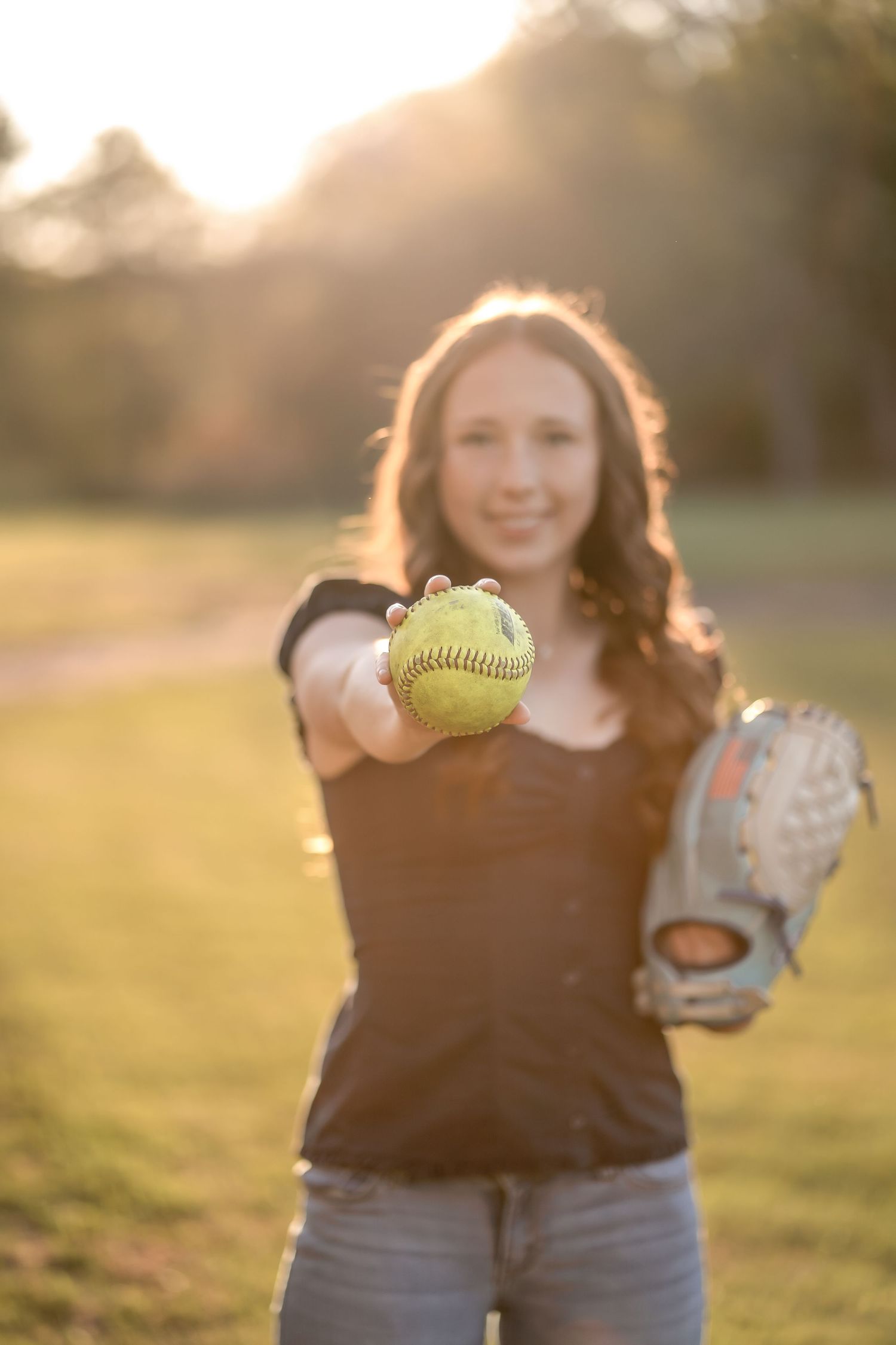 A person in a dark shirt holds up a bright yellow softball against a sunlit outdoor background.