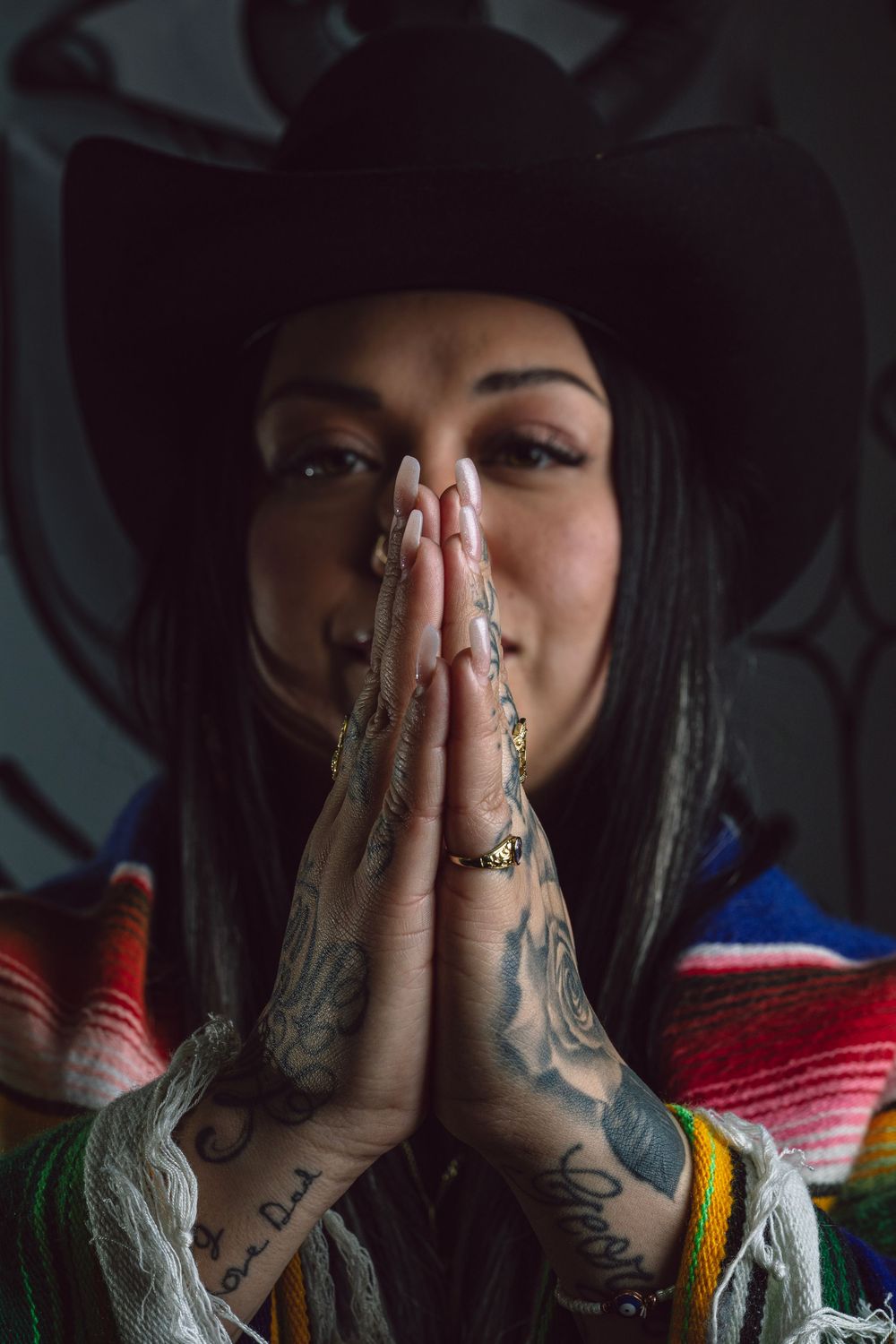 Person wearing a black hat and colorful serape holds tattooed hands in prayer position against dark background.