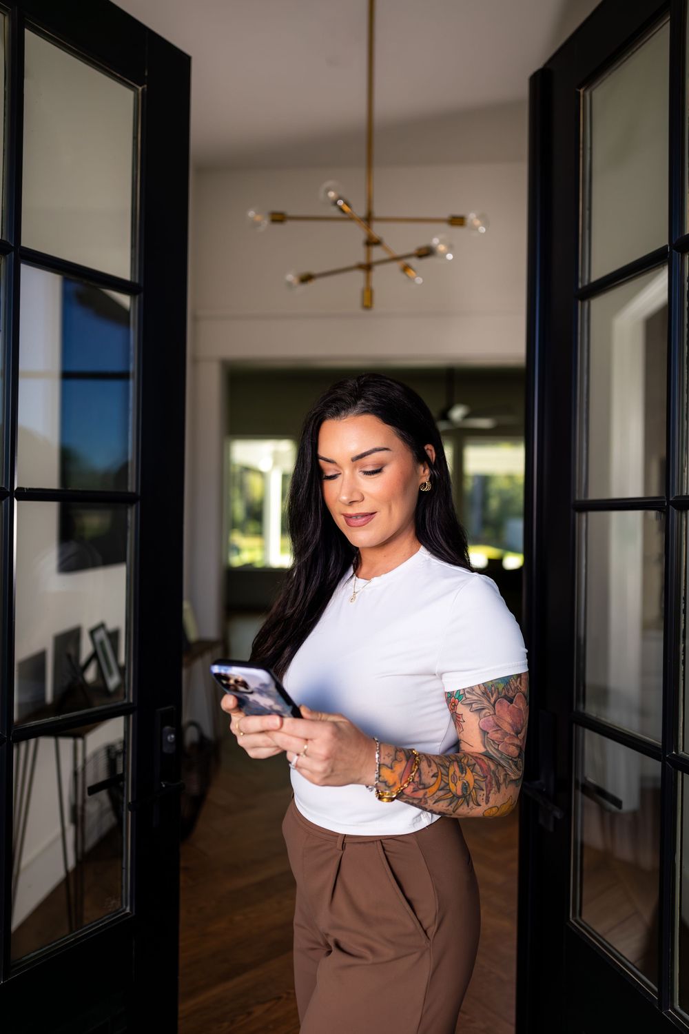 Woman in business casual attire checks phone in modern hallway with black doors.