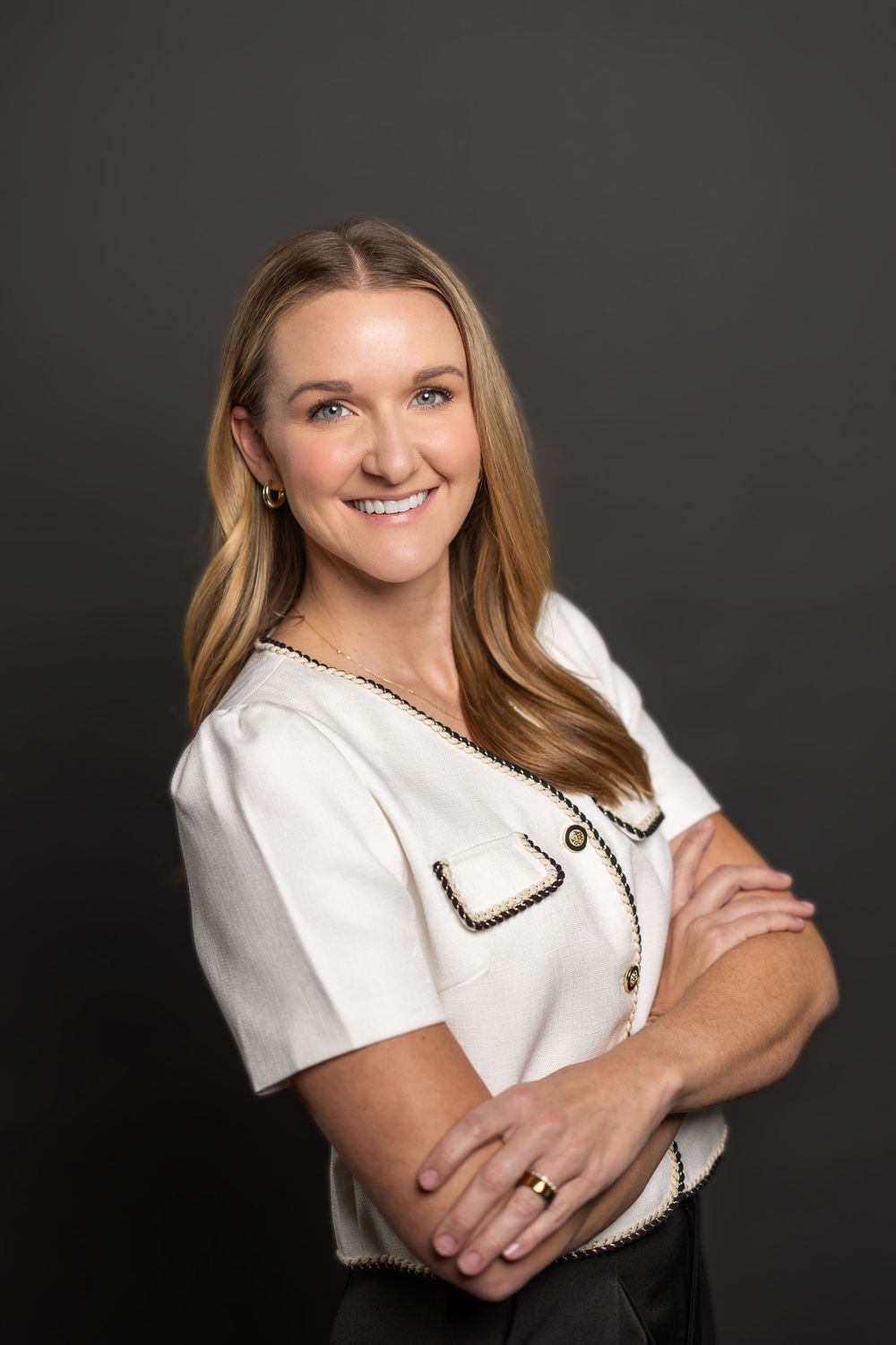 Casual professional portrait with crossed arms wearing white blouse against dark studio backdrop.