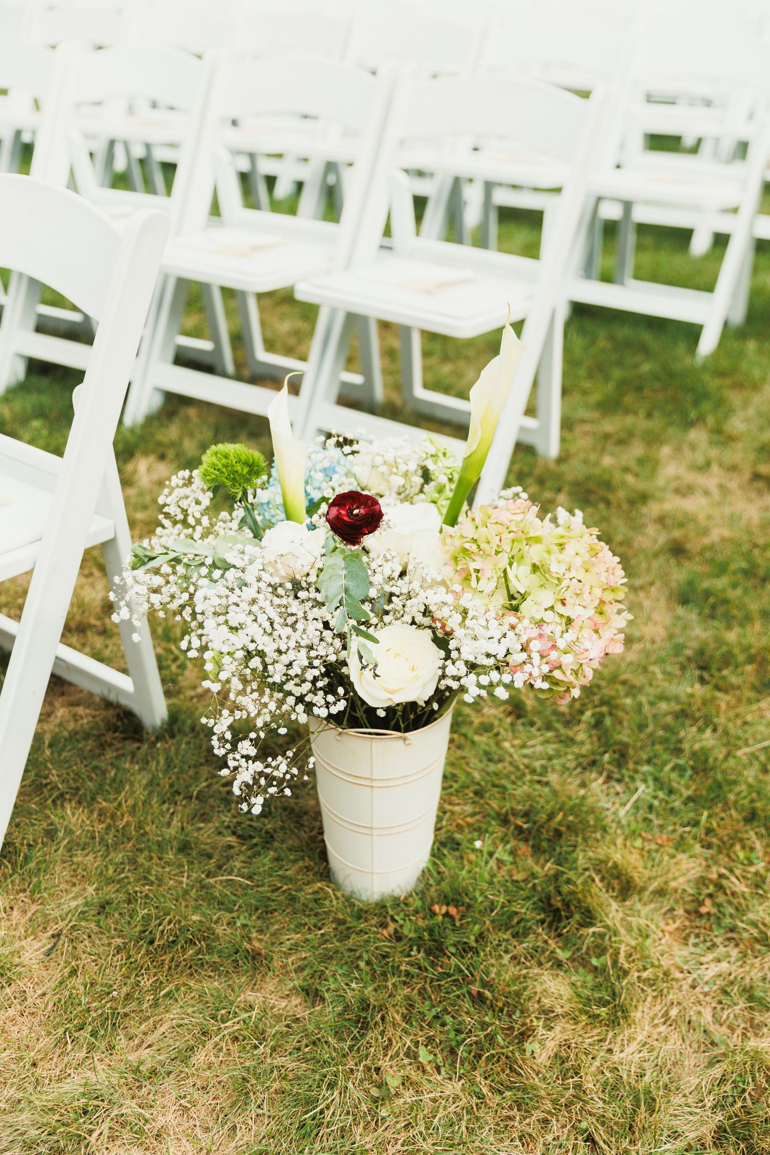 White folding chairs arranged outdoors with small floral bouquets in white buckets along the aisle.