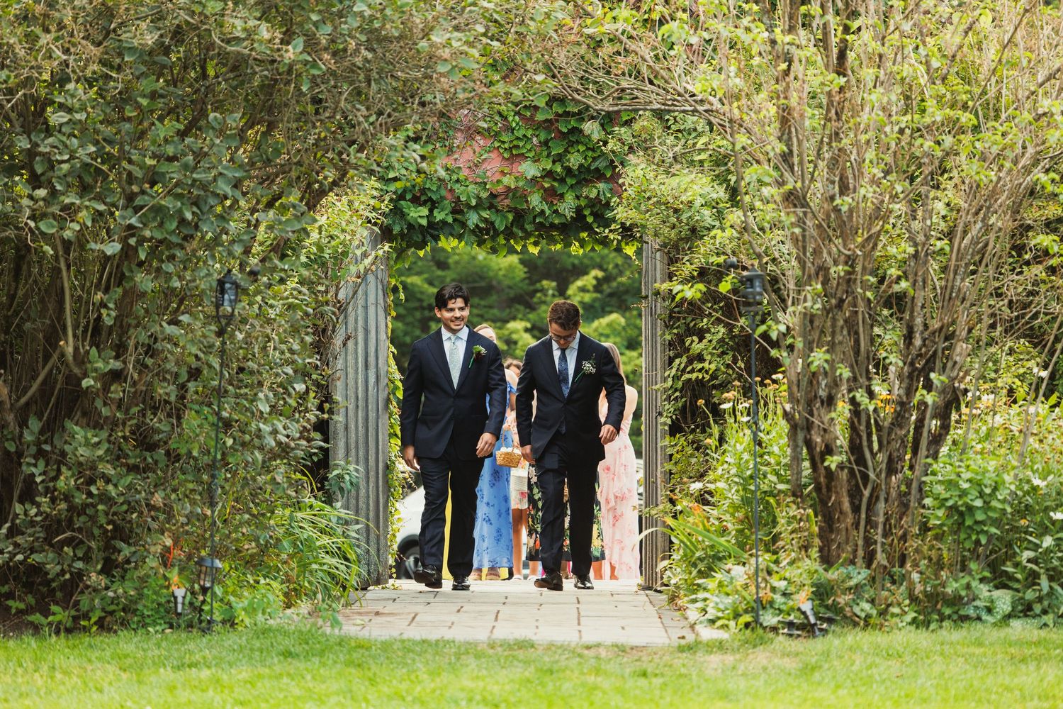 Wedding ceremony processional through a garden pathway lined with trees and flowers.