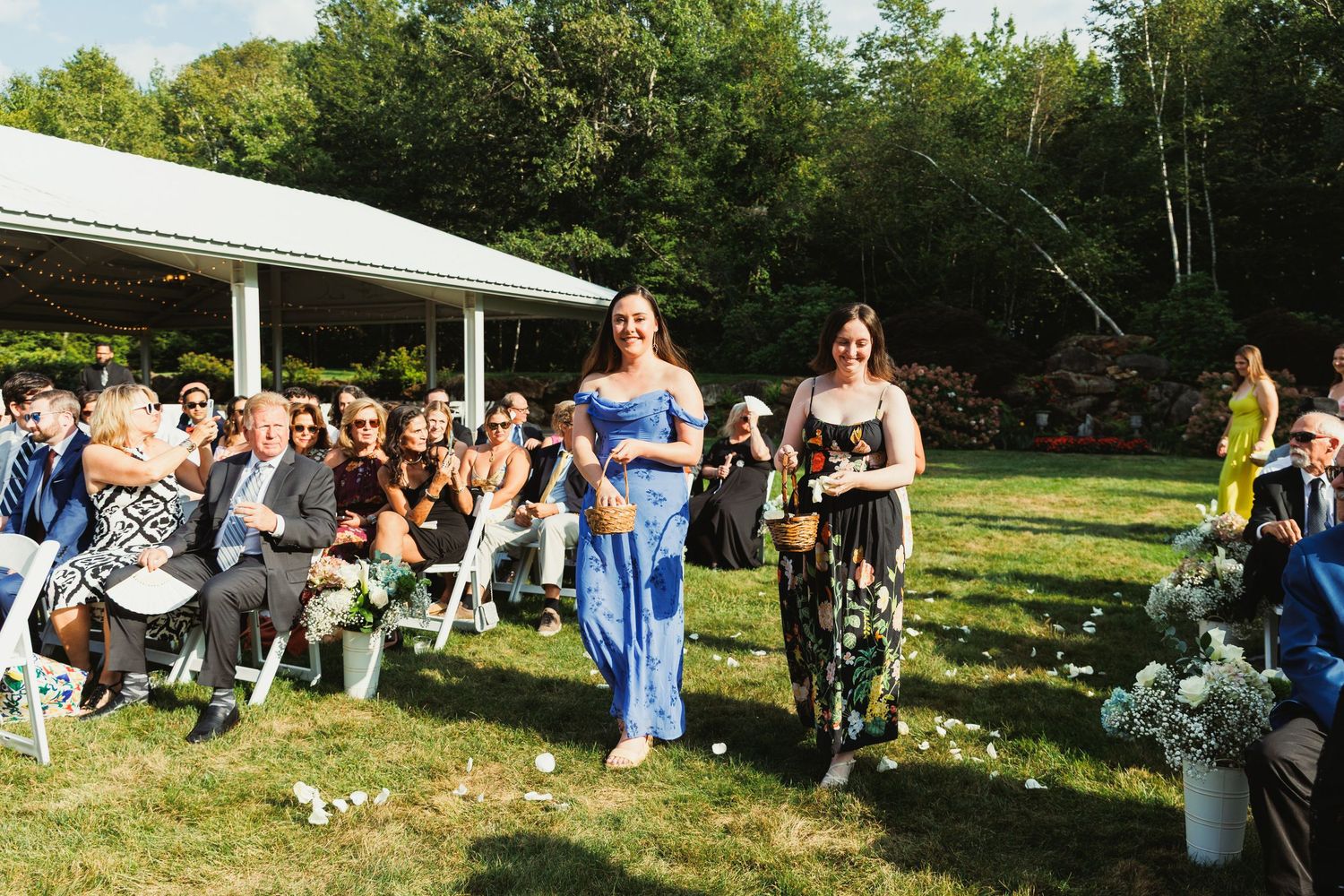 Outdoor wedding reception with guests gathered under a white tent on a sunny day.