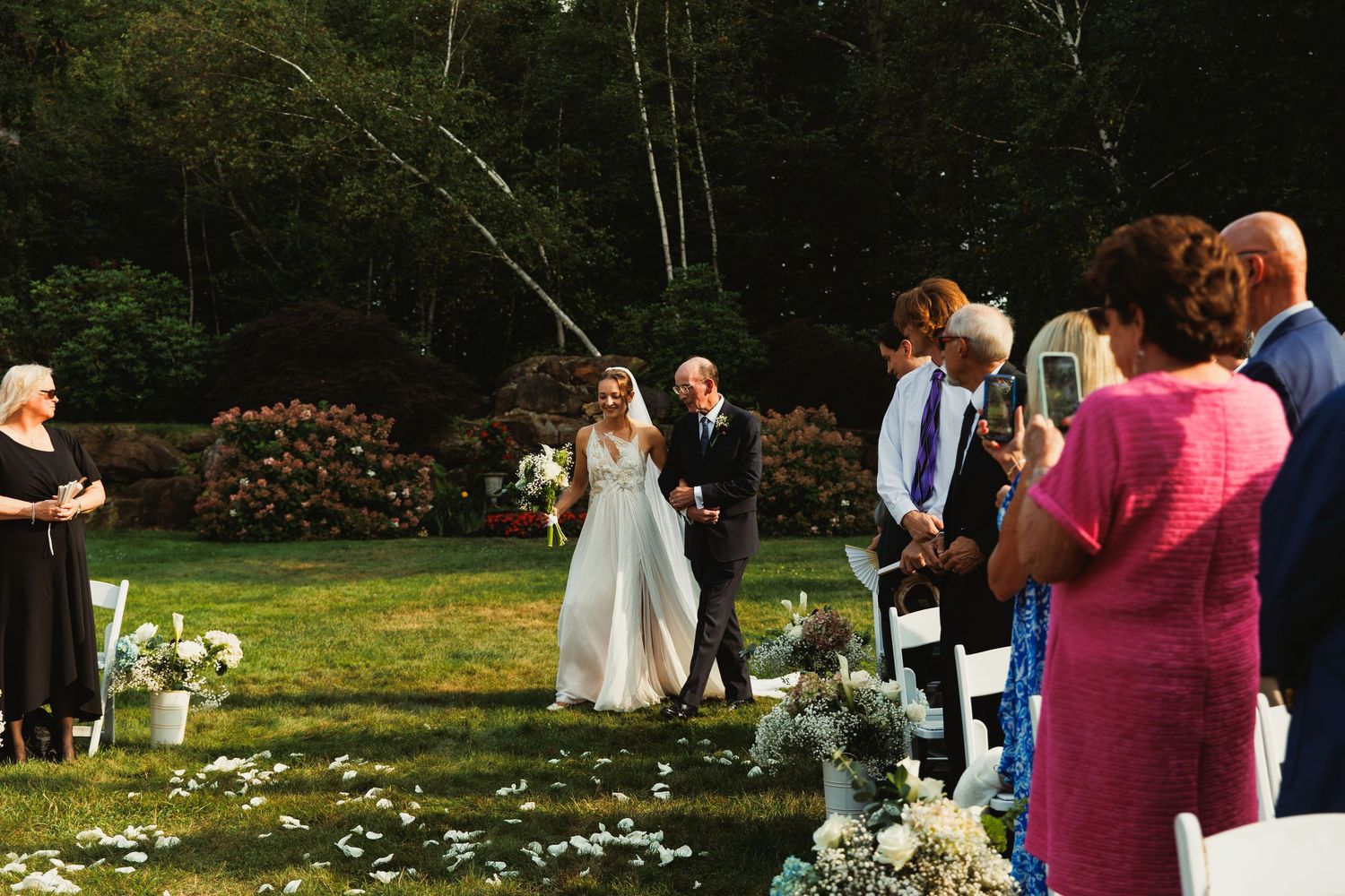 Outdoor wedding ceremony setup with guests seated in chairs on a lawn surrounded by trees.
