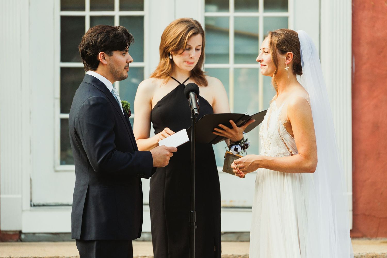 Wedding ceremony participants standing in front of a coral-colored building with white windows.