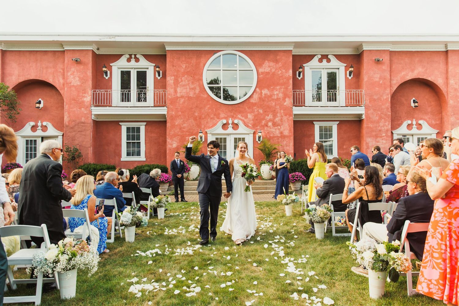 Wedding guests gather on a lawn in front of a historic coral-colored building for an outdoor celebration.