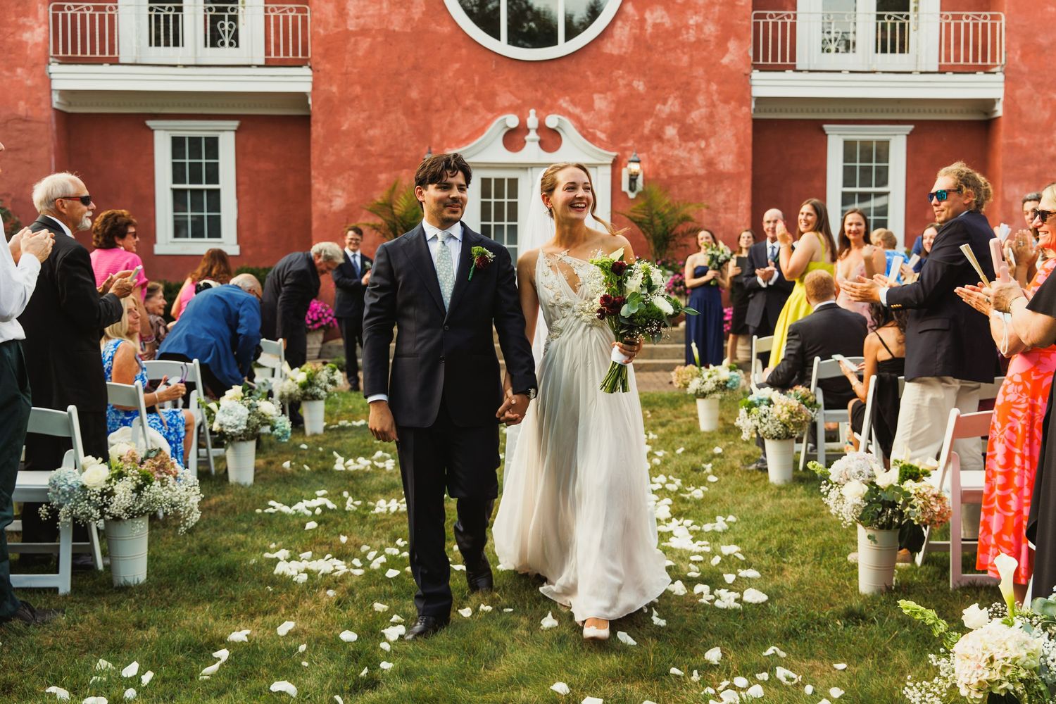 Wedding ceremony setup with white chairs on a lawn in front of a coral-colored mansion.