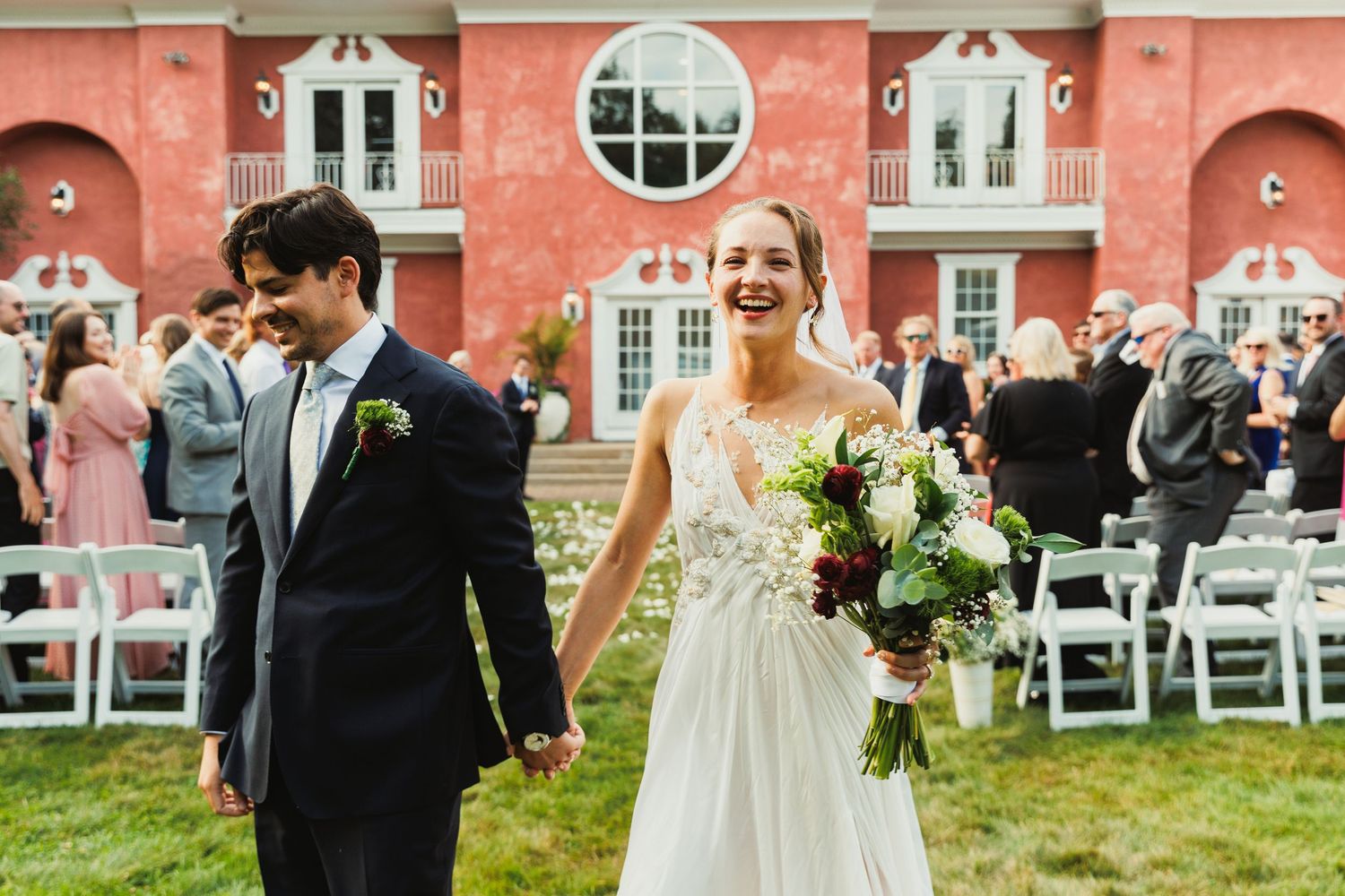Outdoor wedding celebration with guests seated on white chairs against a striking coral building backdrop.