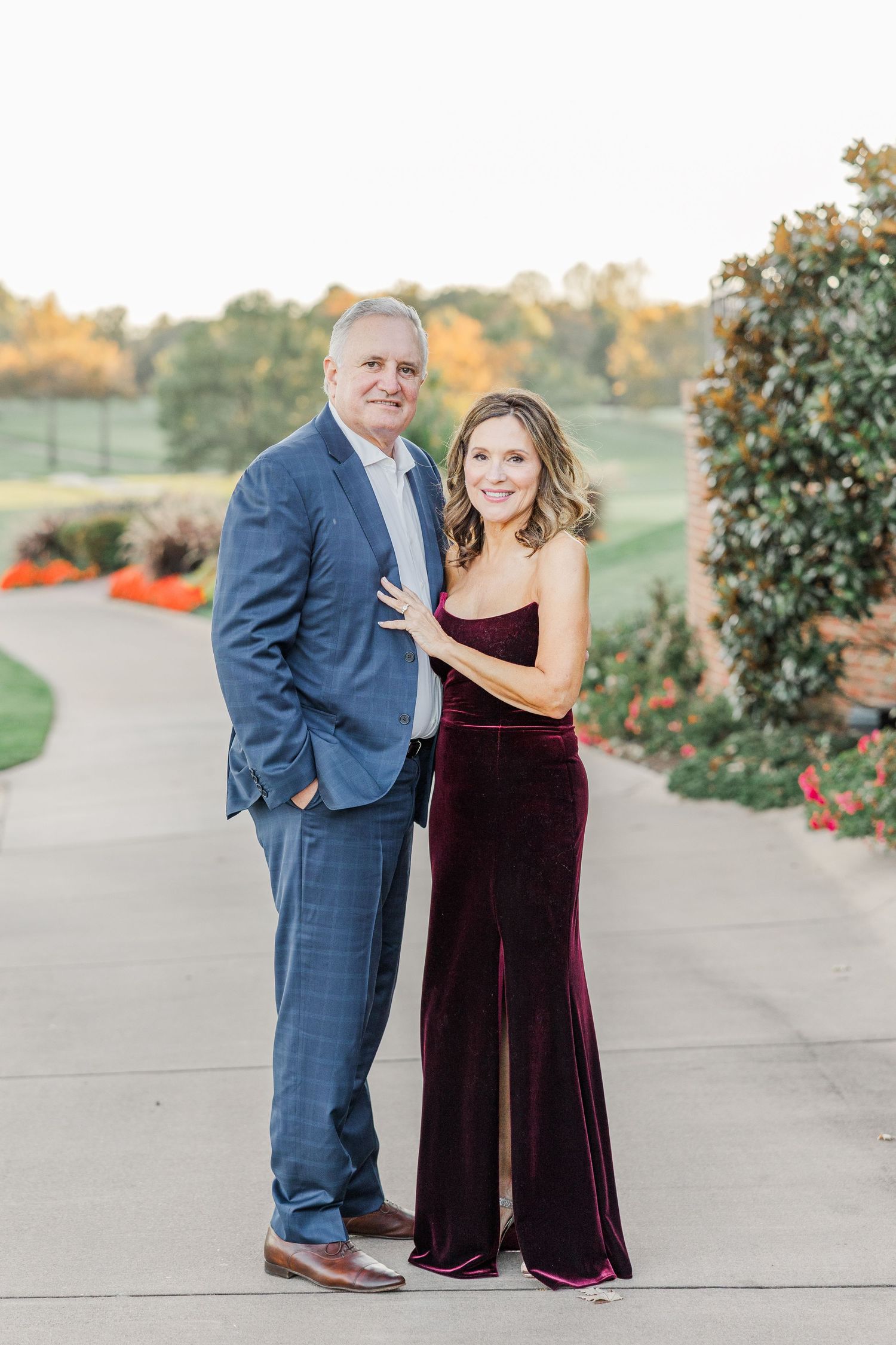 A couple poses outdoors in formal attire, with the woman wearing a burgundy velvet gown and the man in a navy suit.