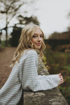 A person in a cozy striped sweater leans against a stone wall outdoors on an overcast autumn day.