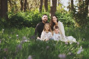 A family sits together on a blanket in a sunlit forest meadow surrounded by wildflowers and tall grass.
