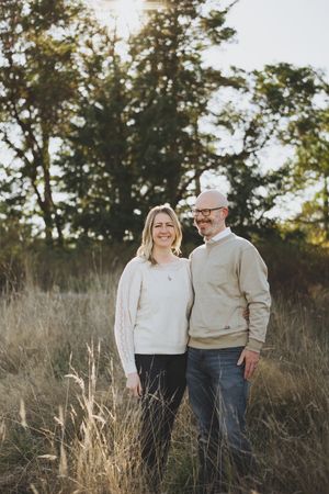 A couple stands together in a sunlit field of tall grass during golden hour.