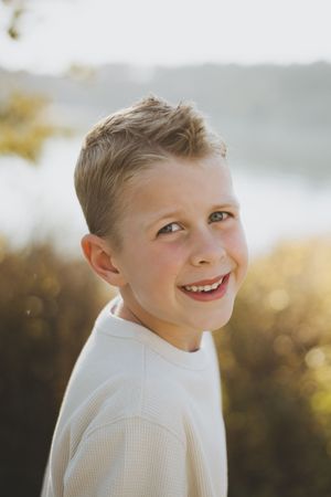 A young child in a white sweater smiles warmly while posing outdoors on a sunny day.