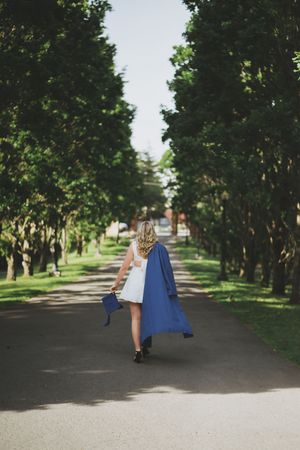 Graduate in blue graduation gown walks down tree-lined path carrying diploma on sunny day.