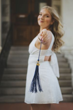 Graduate in white dress holds blue and gold graduation tassel against blurred background.