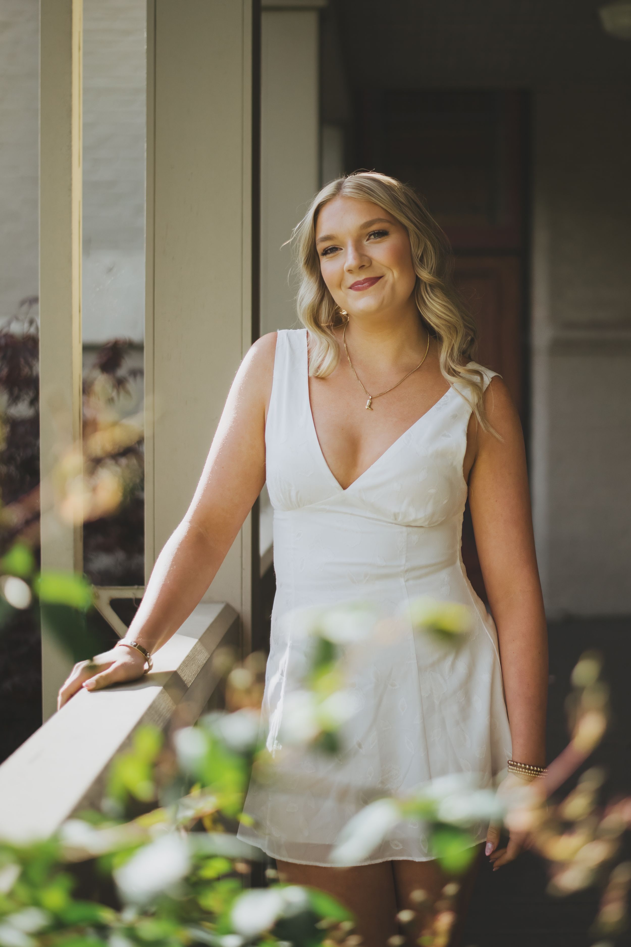 A person in a white sleeveless dress stands on a porch with greenery and natural lighting in the background.