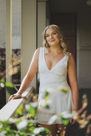 A person in a white sleeveless dress stands on a porch with greenery and natural lighting in the background.