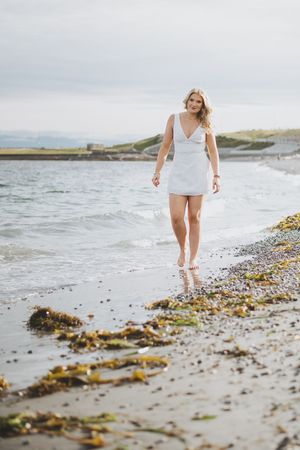 A person in a white dress walks along a beach shoreline covered in seaweed during an overcast day.
