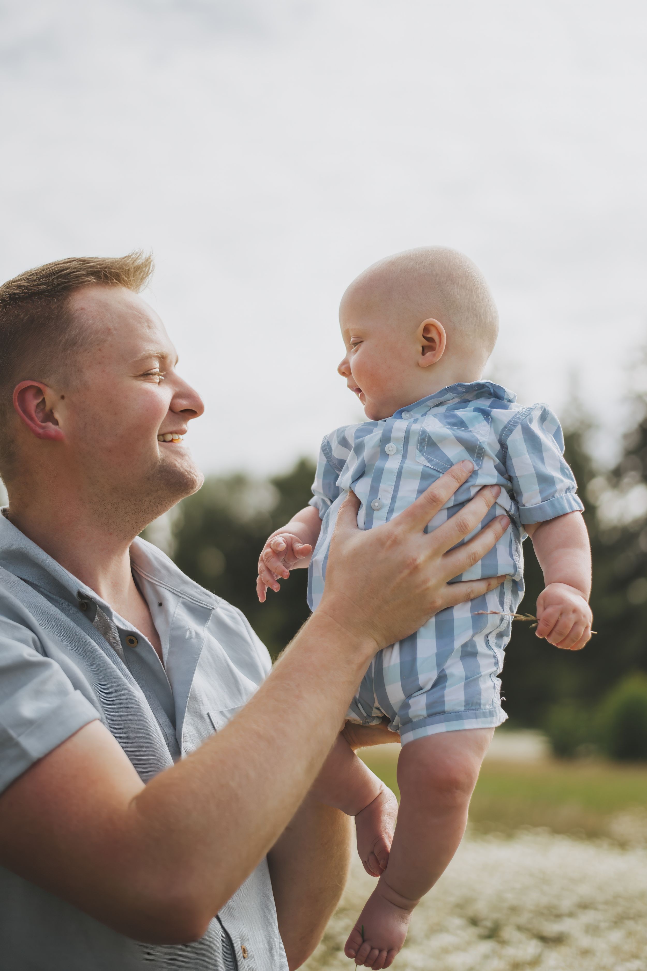 A joyful parent lifts and plays with a small baby wearing blue and white stripes outdoors in nature.