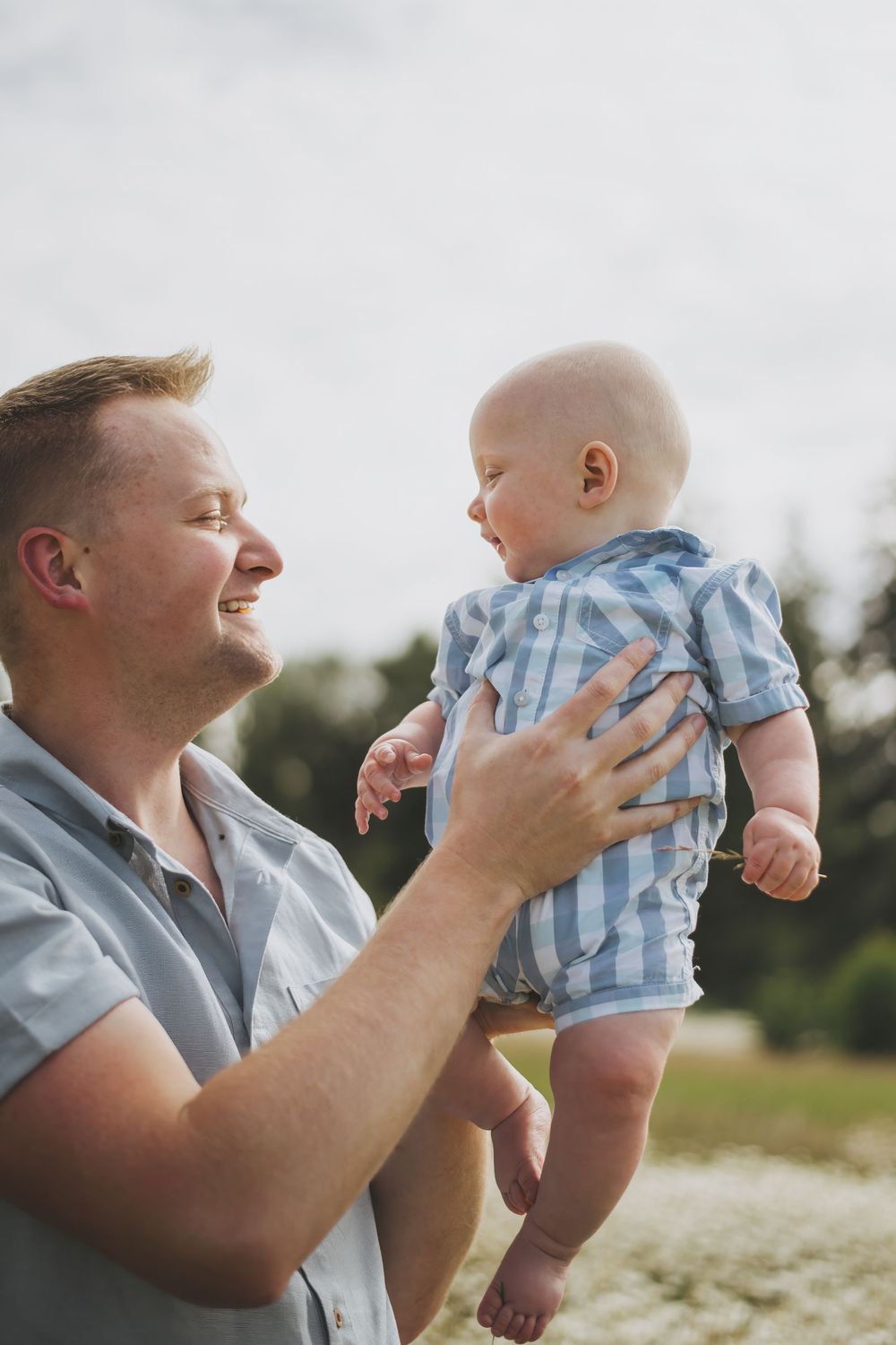 A joyful parent lifts and plays with a small baby wearing blue and white stripes outdoors in nature.