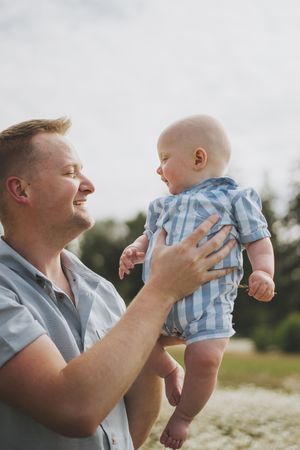 A joyful parent lifts and plays with a small baby wearing blue and white stripes outdoors in nature.