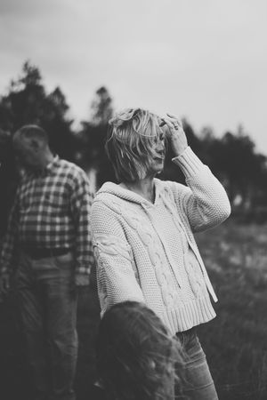 Black and white photo of person in cable knit sweater with hand on head outdoors with blurred figure in background.