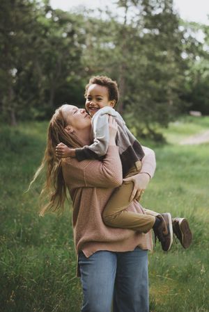 A woman in a pink sweater joyfully lifts a laughing child outdoors in a lush green park setting.
