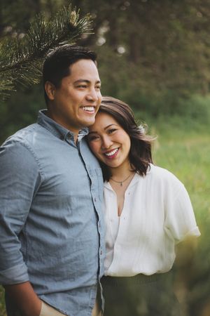 Smiling couple embracing outdoors near pine trees, woman in white blouse resting head on man's shoulder.