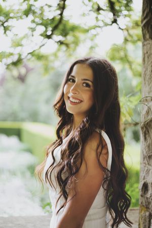 Young woman with long wavy brown hair smiling outdoors in a white dress, surrounded by lush green garden scenery.