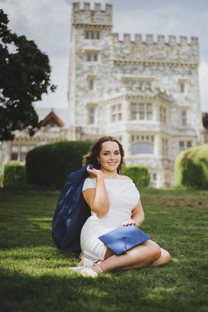 Young woman in white dress sits on grass holding graduation cap and gown before a historic stone castle building.