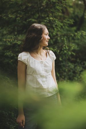 Young woman in white blouse standing peacefully in lush green forest, looking away with a gentle smile.