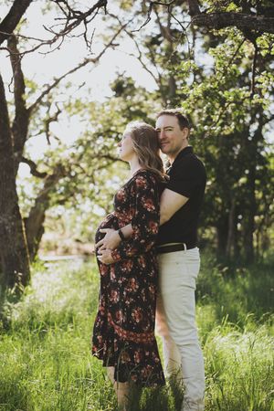 Pregnant couple embracing outdoors in lush green field surrounded by trees during a maternity photo session.