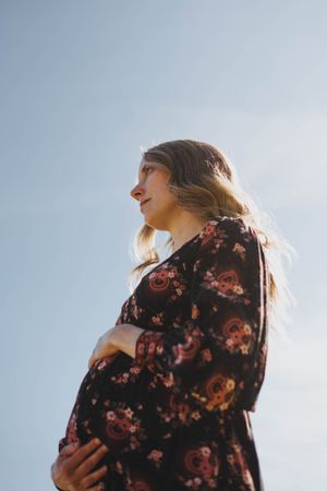 Pregnant woman in floral dress cradling baby bump outdoors against blue sky, glowing in natural sunlight.