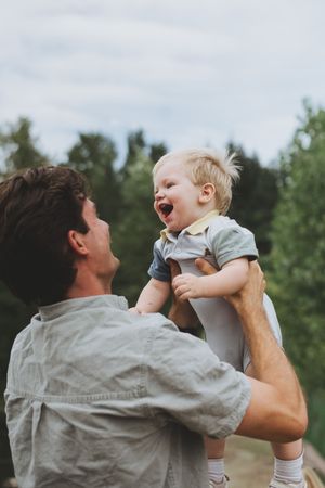 A joyful father lifts his laughing blonde toddler outdoors, sharing a playful moment against a lush green tree backdrop.