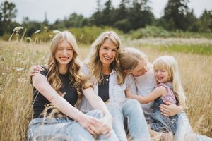 Four smiling females sitting together in a sunny meadow with wildflowers, enjoying a cheerful outdoor family moment.